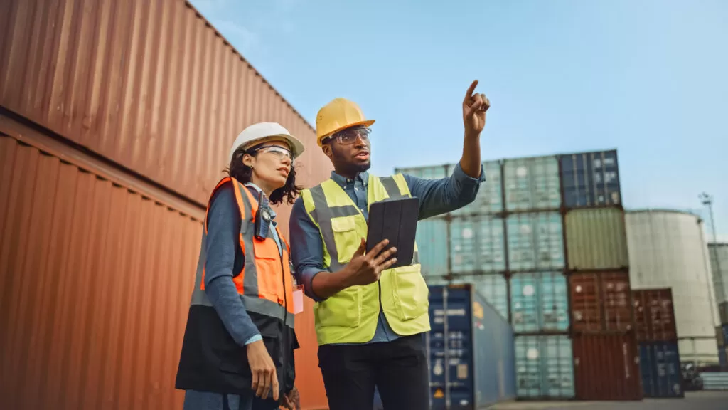 Multiethnic Female Industrial Engineer with Tablet and Black African American Male Supervisor in Hard Hats and Safety Vests Stand in Container Terminal. Colleagues Talk About Logistics Operations.